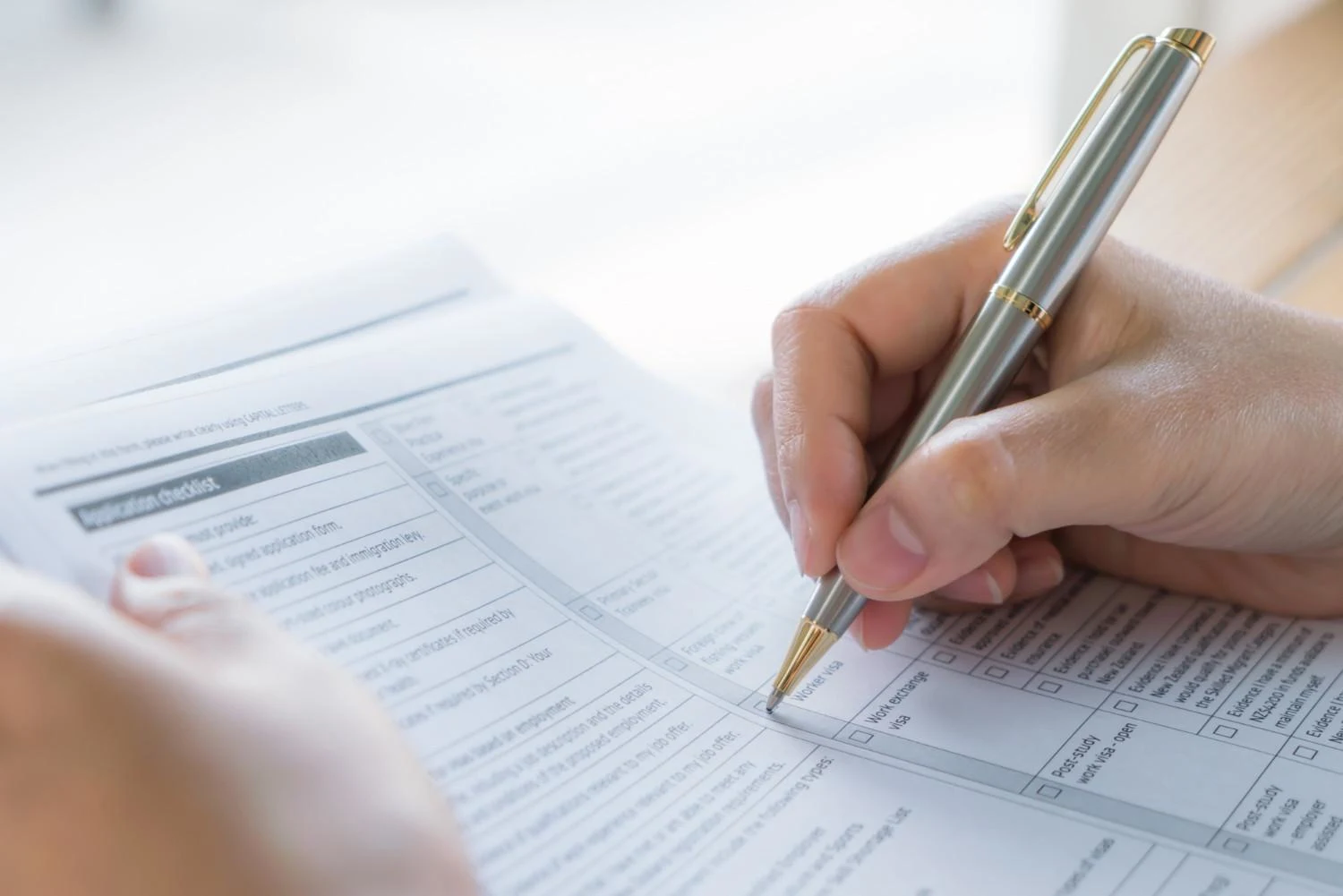 A hand holding a silver pen fills out a checklist on a paper form. The focus is on the pen and hand, conveying concentration and attention to detail.